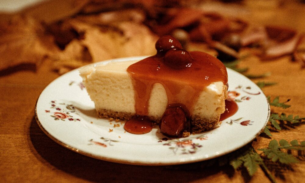 Na slici je komad torte preliven karamel umakom, poslužen na tanjuru s cvjetnim uzorkom. Torta je bogato prekrivena umakom. Pozadina slike prikazuje stolnjak s listopadnim uzorkom, dajući toplu i ugodnu atmosferu slici.