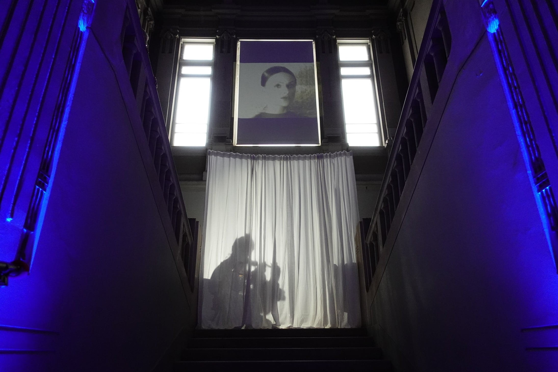 Ova slika prikazuje stubište osvijetljeno plavim svjetlom s bijelim zastorima koji prekrivaju prozor na vrhu. Na zastorima se vidi sjena osobe. Stubište je obasjano plavim svjetlima duž zidova, stvarajući mračnu i dramatičnu atmosferu. Iznad zastora nalazi se umjetničko djelo.