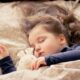 Na slici se vidi djevojčica koja leži na krevetu s uzorkom cvjetova. Djevojčia drži plišanu igračku koja izgleda kao zec ili medvjed. Atmosfera na slici je mirna i opuštena.