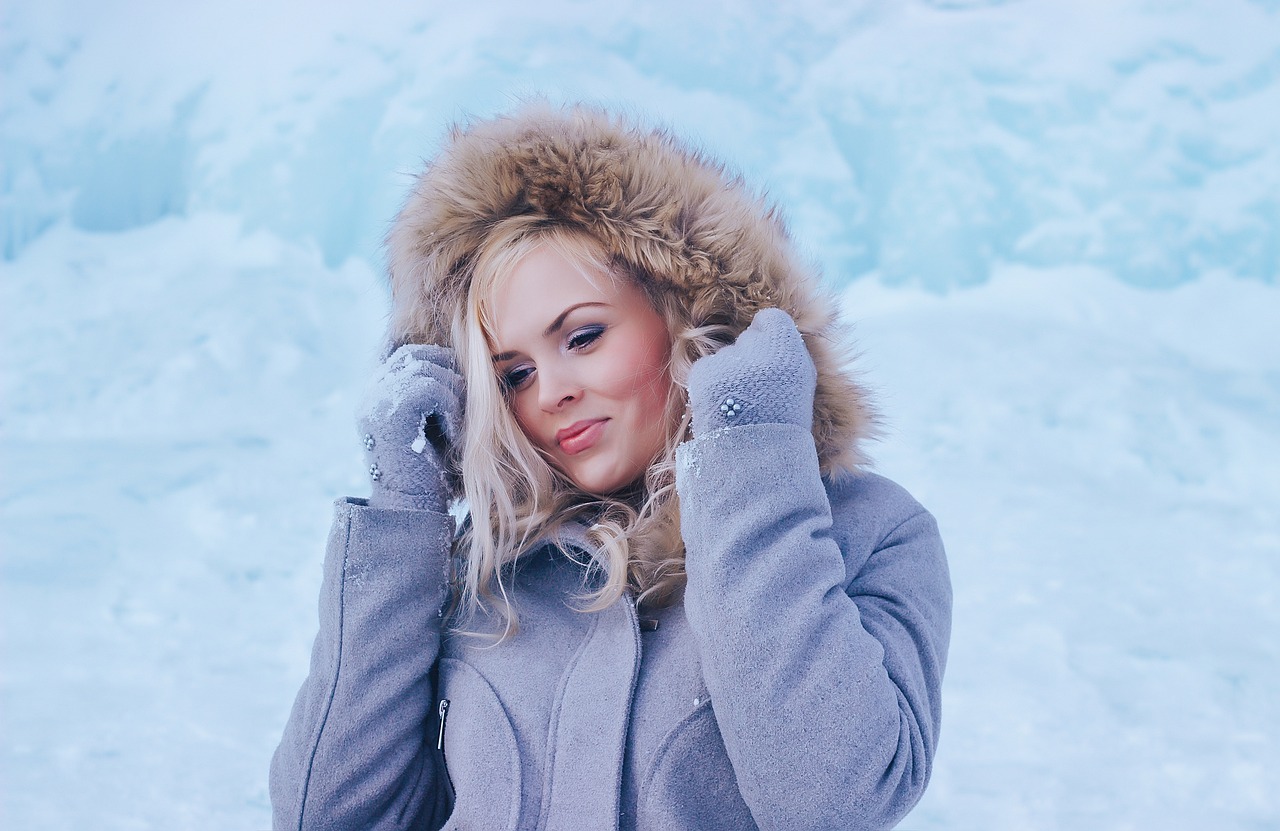 Na slici je osoba u zimskom okruženju, okružena snijegom i ledom. Osoba nosi kaput s kapuljačom obloženom krznom, koja je podignuta preko glave. Rukavice su joj prekrivene snijegom, a drži ih blizu lica.