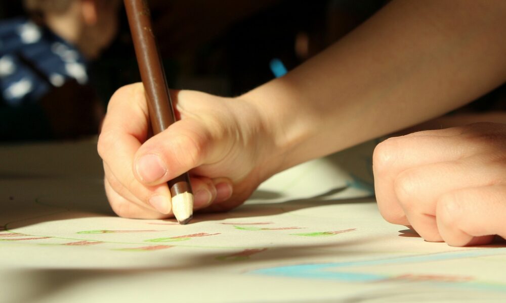 Na fotografiji se vidi ruka koja drži olovku i crta na papiru. Papir je prekriven raznobojnim crtama koje izgledaju kao dio dječjeg crteža. Ruke su male i delikatne, što sugerira da pripadaju djetetu. Pozadina je zamućena, ali se naziru elementi unutarnjeg prostora s prirodnom svjetlošću. Naglasak je na ruci koja drži olovku, prikazujući čin crtanja.