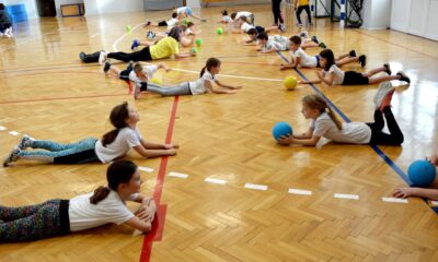 Ova slika prikazuje grupu djece koja vježba u sportskoj dvorani. Djeca su raspoređena po podu i drže lopte raznih boja. Pod je drven, sa crvenim i plavim crtama. Na zidovima dvorane vidljivi su svjetlosni reflektori, a pod je sjajno poliran.
