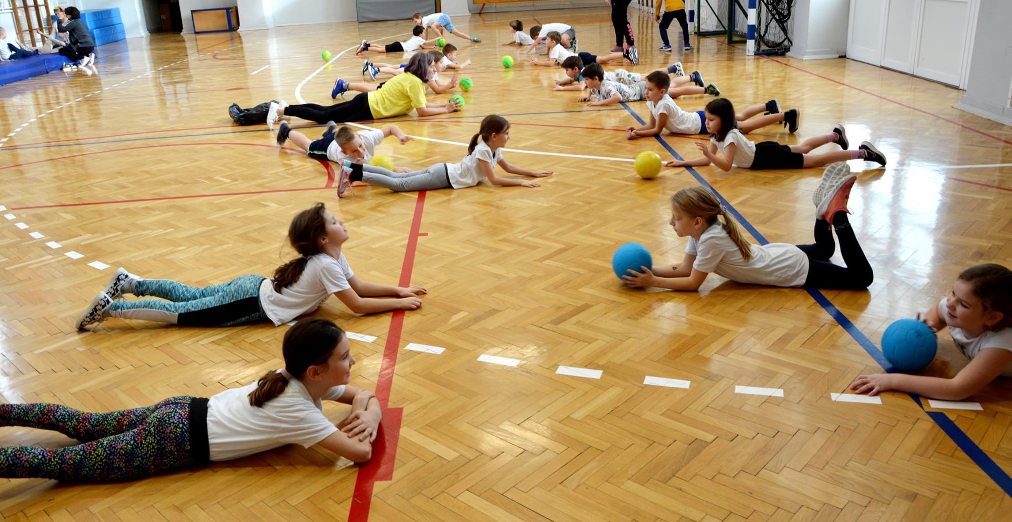 Ova slika prikazuje grupu djece koja vježba u sportskoj dvorani. Djeca su raspoređena po podu i drže lopte raznih boja. Pod je drven, sa crvenim i plavim crtama. Na zidovima dvorane vidljivi su svjetlosni reflektori, a pod je sjajno poliran.