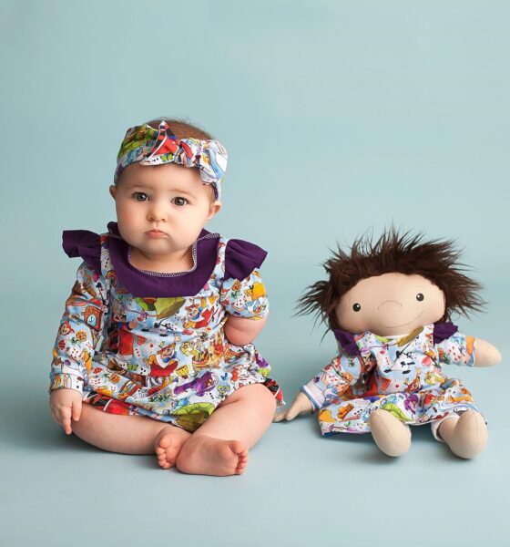 Na fotografiji je beba koja sjedi pored lutke na plavoj pozadini. Beba i lutka nose identične haljinice s šarenim printom.
