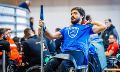 Na slici je osoba u plavoj majici koja sjedi u invalidskim kolicima i drži kuglu štap. Osoba je okružena drugim ljudima koji također sjede u invalidskim kolicima. Svi su unutar sportske dvorane,