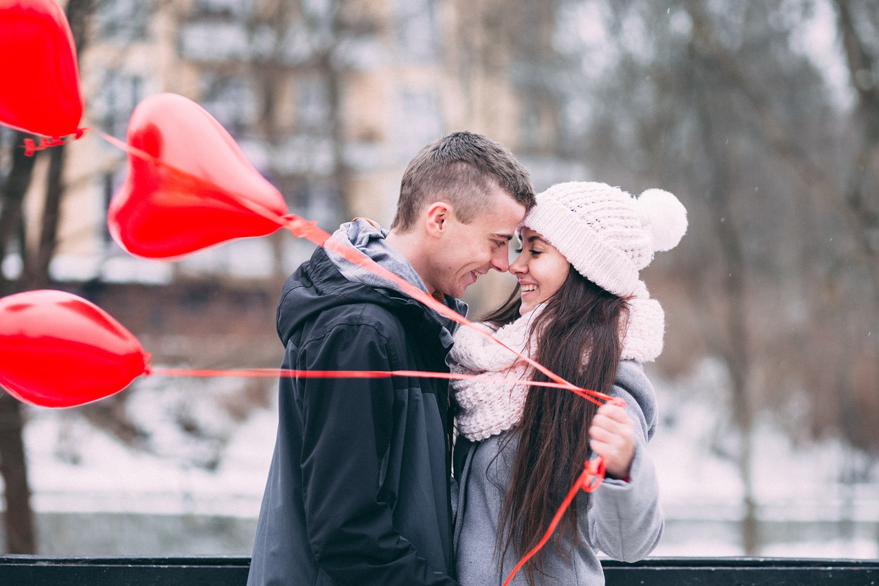 Fotografija prikazuje dvije osobe koje se grle i drže crvene balone u obliku srca. Pozadina slike pokazuje snijegom prekriveno područje, što sugerira da je zima.