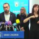 Fotografija prikazuje skupinu ljudi koji stoje iza plavog stola s mikrofonima. Lica su im zamagljena. Na stolu je postavljeno nekoliko mikrofona različitih medijskih kuća. Pozadina sadrži logotip i tekst “GRAD ZAGREB”.