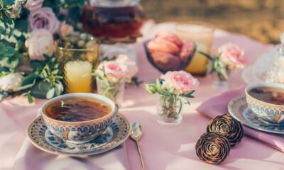 Fotografija prikazuje stol postavljen za čajanku na otvorenom. Na stolu prekrivenom ružičastim stolnjakom nalaze se šalice s čajem, poslužavnici, vaza s ružama i drugi cvjetni aranžmani. Sve je osvijetljeno prirodnom svjetlošću koja dodaje toplinu sceni.