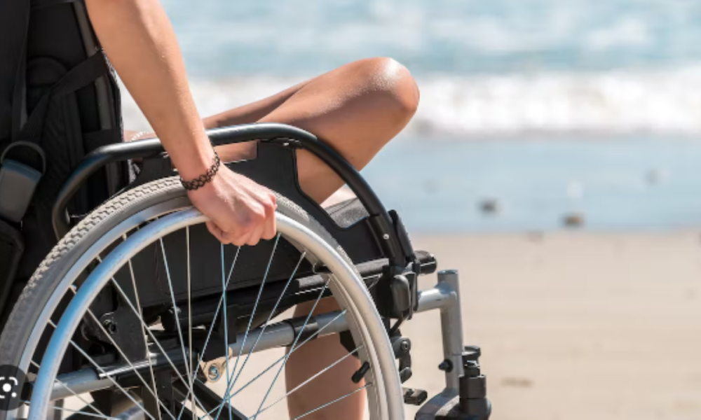 Na fotografiji je osoba koja sjedi u invalidskim kolicima na plaži. Fokus je na ruci osobe koja drži kotač kolica, a pozadina prikazuje more i pijesak. Osoba nosi crnu odjeću i ima narukvicu na zapešću. Invalidska kolica imaju crni jastuk i metalnu konstrukciju s plavim detaljima na kotaču. U pozadini se pruža pogled na pješčanu plažu s valovima koji se razbijaju pod jakim sunčevim svjetlom.