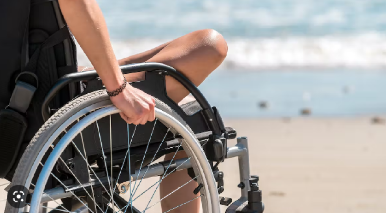 Na fotografiji je osoba koja sjedi u invalidskim kolicima na plaži. Fokus je na ruci osobe koja drži kotač kolica, a pozadina prikazuje more i pijesak. Osoba nosi crnu odjeću i ima narukvicu na zapešću. Invalidska kolica imaju crni jastuk i metalnu konstrukciju s plavim detaljima na kotaču. U pozadini se pruža pogled na pješčanu plažu s valovima koji se razbijaju pod jakim sunčevim svjetlom.