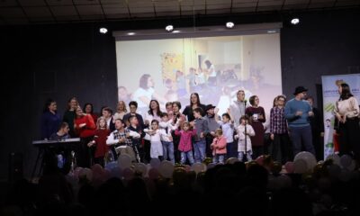 Fotografija prikazuje grupu ljudi na pozornici, s projekcijom u pozadini. Na sceni je raznolika skupina ljudi, uključujući djecu i odrasle. Pozadinska projekcija prikazuje sliku sobe s ljudima.