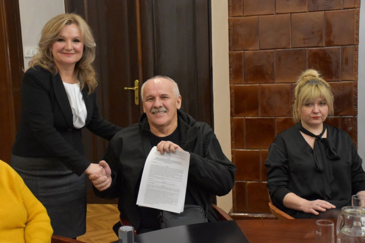 Na fotografiji su tri osobe. Osoba u sredini, obučena u crnu odjeću, drži papir i rukuje se s osobom s lijeve strane koja je obučena u bijelu bluzu i crni sako. Osoba s desne strane, također obučena u crno, sjedi za stolom. Pozadina prikazuje drvena vrata i zid.