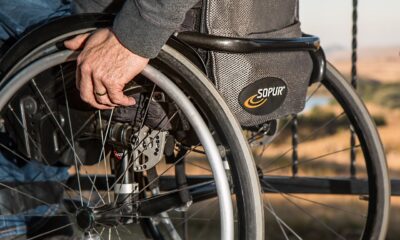 Fotografija prikazuje osobu koja sjedi u invalidskim kolicima. Fokus je na ruci osobe i detaljima invalidskih kolica, uključujući kotače i kočnice. Pozadina slike prikazuje otvoreno polje, ali nije jasno vidljiva zbog fokusa na predmete u prvom planu. Invalidska kolica su označena brendom “SOPUR”.