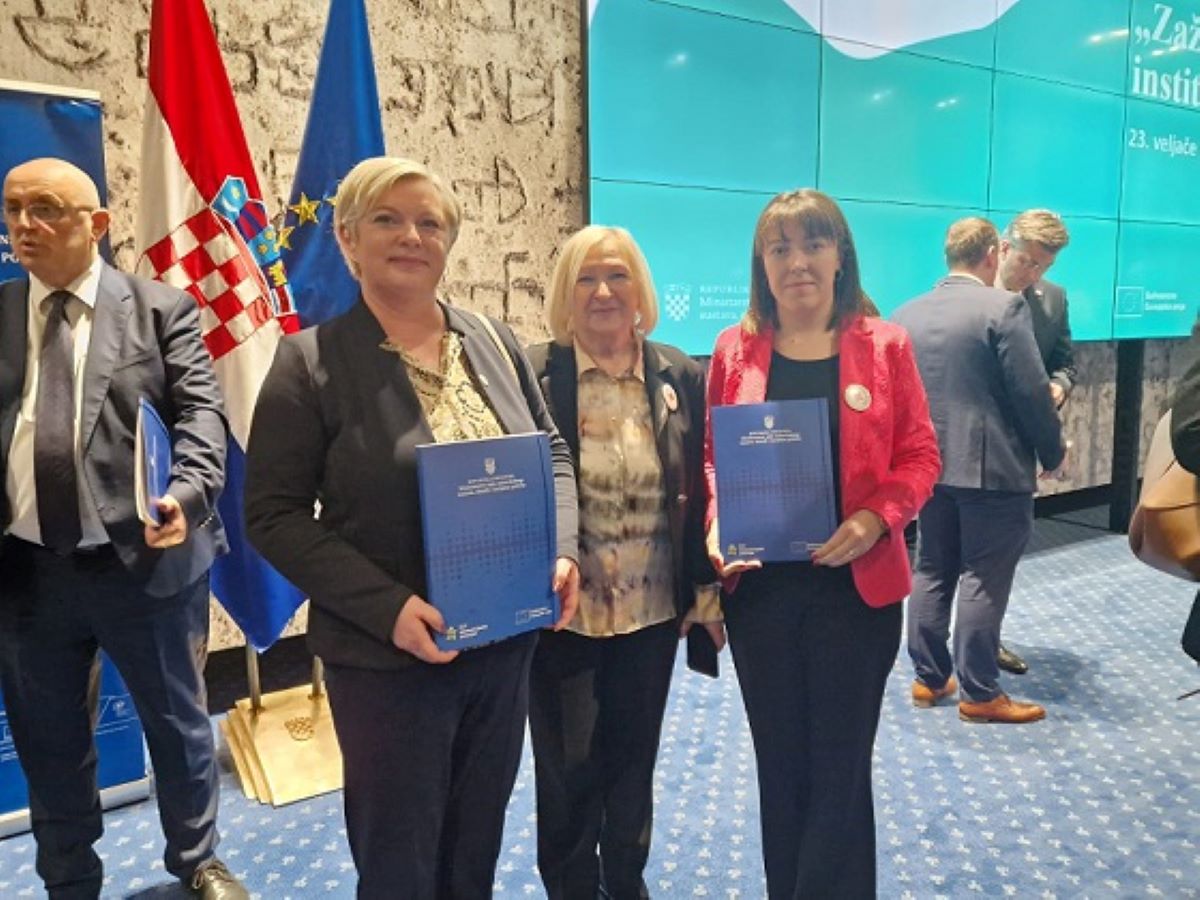 Na fotografiji su četiri osobe. Stoje ispred plave pozadine i drže plave mape. Iza njih su zastave Europske unije i Hrvatske. Osobe su odjevene formalno. Fotografija sugerira da se radi o službenom ili formalnom događaju.