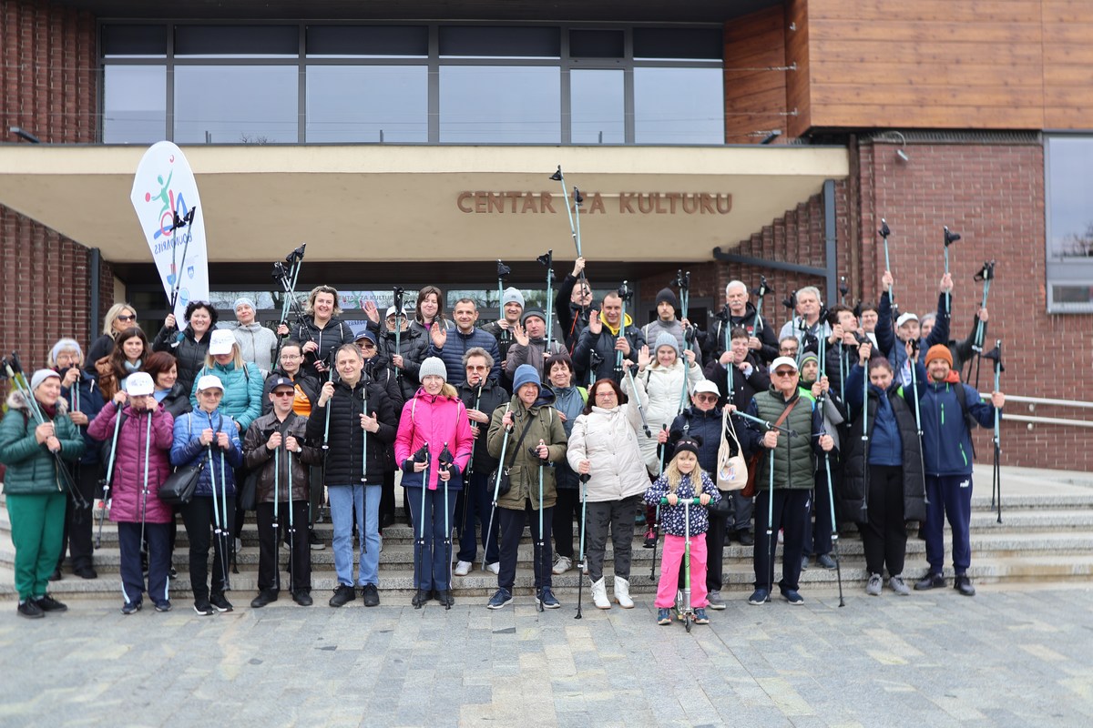 Fotografija prikazuje grupu ljudi koji drže štapove za hodanje ispred zgrade s natpisom 'CENTAR ZA KULTURU'. Grupa se sastoji od osoba različitih dobnih skupina, uključujući djecu i odrasle. Zgrada je modernog dizajna s velikim staklenim prozorima i vratima. Na lijevoj strani slike nalazi se zastava ili transparent s nečitkim tekstom zbog kuta i udaljenosti.