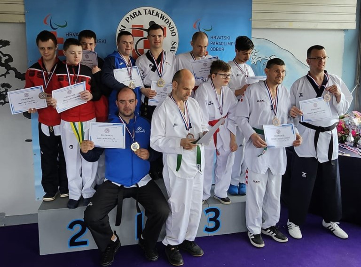 Na slici je grupa ljudi koji su osvojili nagrade na natjecanju u parataekwondou. svi drže diplome u rukama. Pozadina prikazuje plakat sa natpisom “PARA TAEKWONDO” Na podiju su označena mjesta 1, 2 i 3, što ukazuje na pobjedničke pozicije.