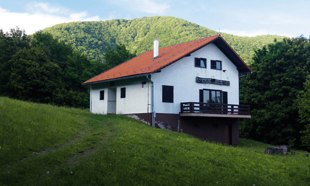 Ova slika prikazuje dvokatnu kuću s bijelom fasadom i tamnim detaljima, smještenu u zelenom okruženju. Kuća ima crveni krov i prostranu terasu na gornjem katu. Staza vodi prema kući, a oko nje je travnata površina. U pozadini se proteže gusta šuma koja vodi prema planini.