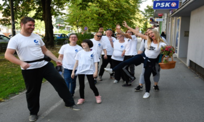 Na fotografiji je grupa ljudi koja nosi bijele majice s plavim logom. Hodaju pločnikom pored zgrade. Okolina izgleda urbano i zeleno drveće je vidljivo u pozadini. Žena s desne strane slika nosi košaru punu cvijeća.