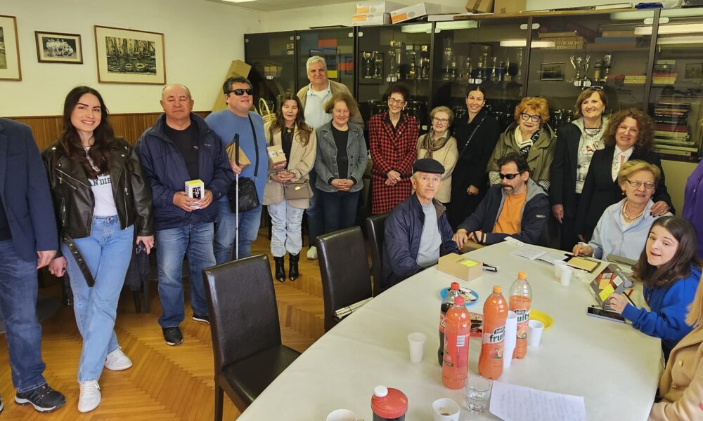 Na fotografiji je grupa ljudi u Udruzi slijepih Split. Na stolu se nalaze različiti predmeti, uključujući papire i boce narančinog soka. Osobe su obučene u ležernu odjeću, uključujući jakne i traperice. U pozadini se vidi prozor koji dopušta prirodno svjetlo u prostoriju.