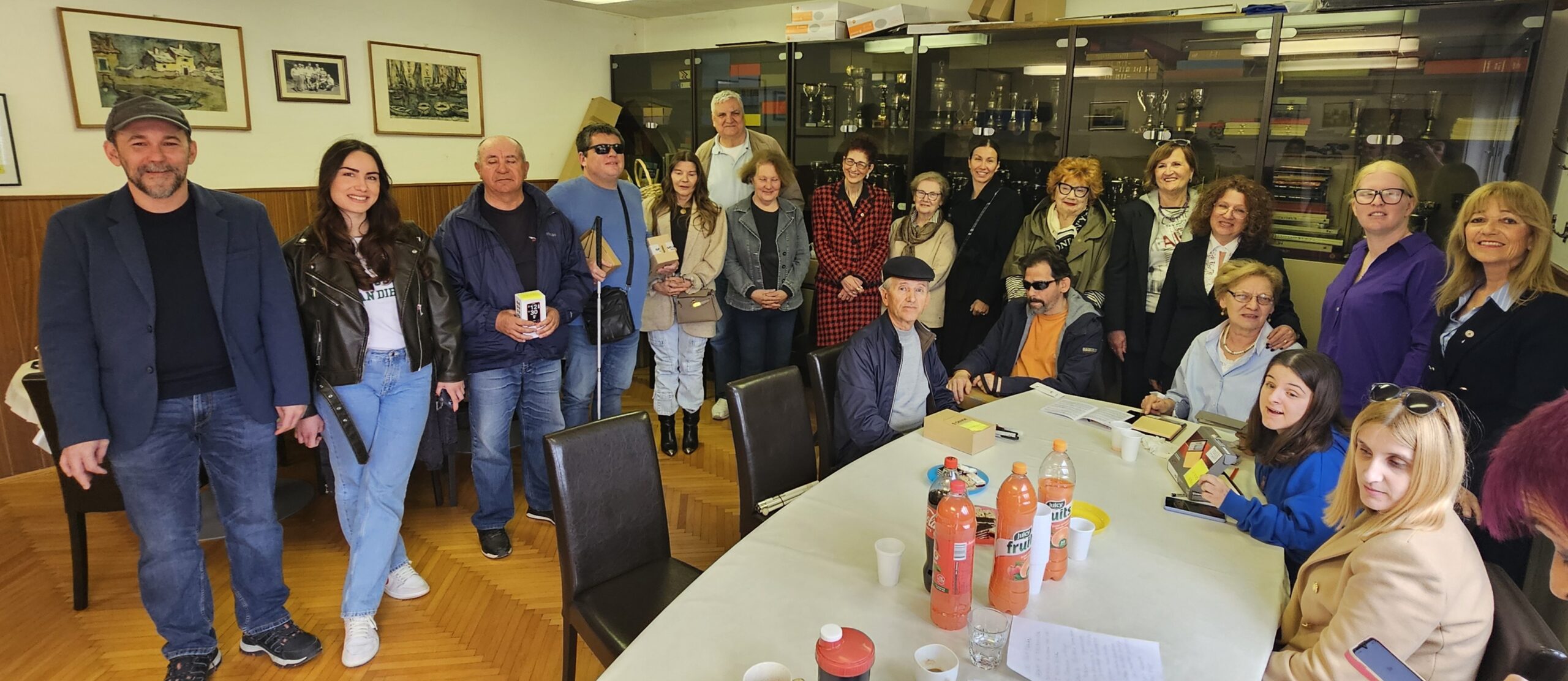 Na fotografiji je grupa ljudi u Udruzi slijepih Split. Na stolu se nalaze različiti predmeti, uključujući papire i boce narančinog soka. Osobe su obučene u ležernu odjeću, uključujući jakne i traperice. U pozadini se vidi prozor koji dopušta prirodno svjetlo u prostoriju.