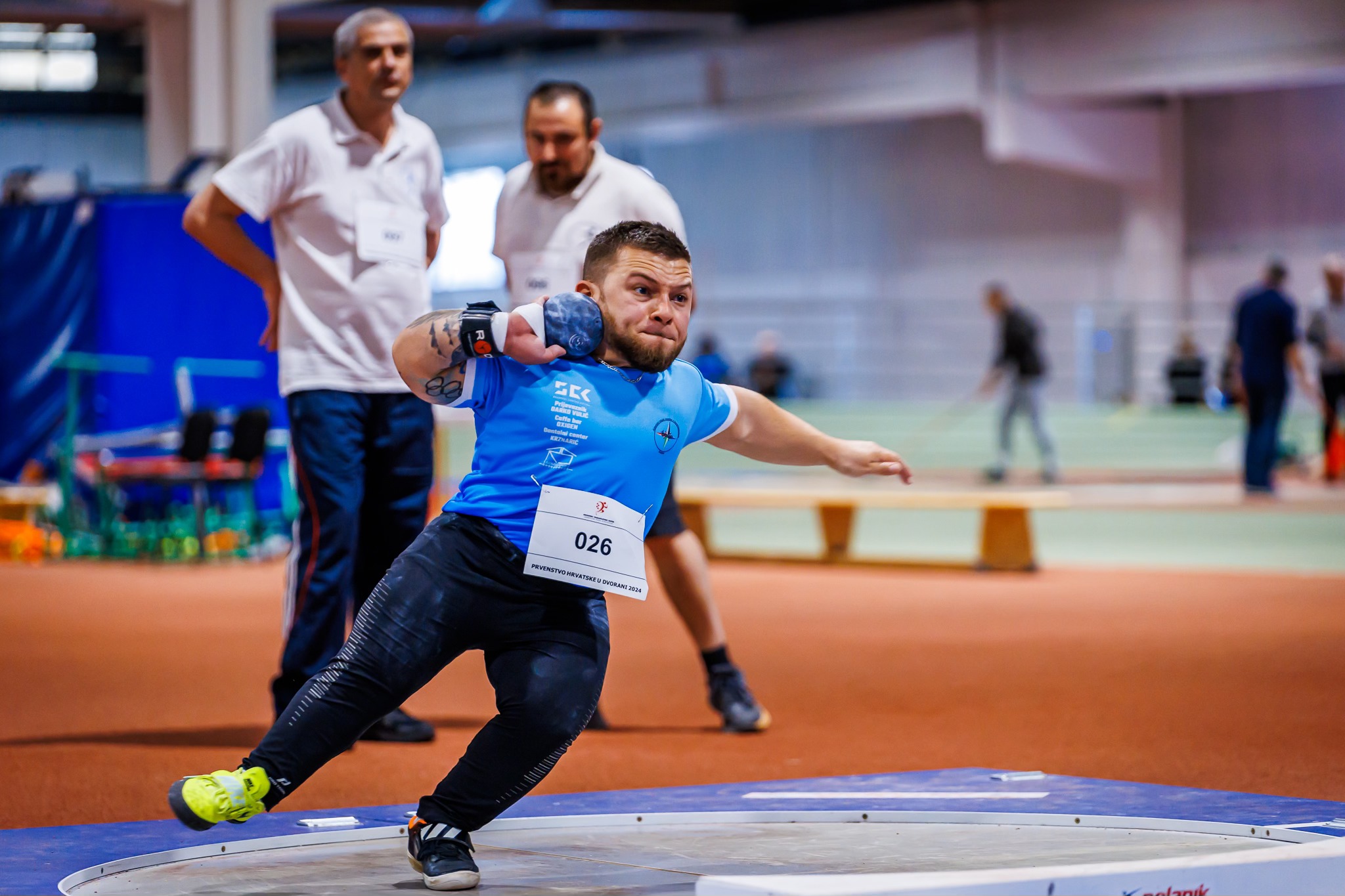 Na slici je atletičar koji se priprema baciti kuglu. Radi se o nekoj vrsti atletskog natjecanja u dvorani. Natjecatelj ima plavu majicu, na njoj je redni broj natjecatelja i naslov "Hrvatski paraatletski savez.