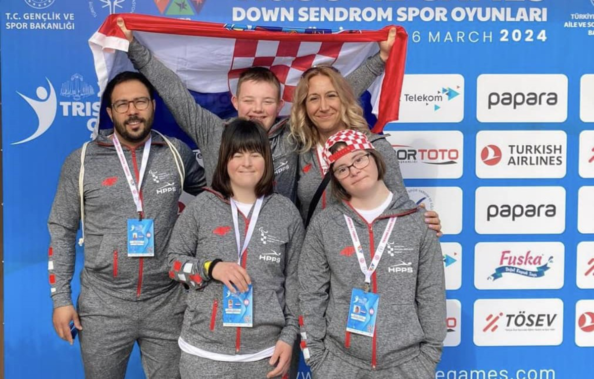 Na slici je grupa ljudi koji nose sive trenirke i medalje. Stoje ispred plavog banera na kojem pišu informacije o sportskom događaju za osobe s Down sindromom. Drže hrvatsku zastavu koja ima crvene, bijele i plave boje. Na baneru se nalaze logotipi sponzora kao što su Turkish Airlines i papara, među ostalima. Događaj je povezan s sportskim aktivnostima za osobe s Down sindromom.