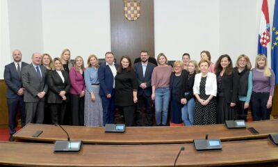 Na fotografiji je grupa ljudi koja stoji u prostoriji s državnim grbom i zastavama u pozadini. Ispred njih je dugi stol s mikrofonima. Ova postavka sugerira da se radi o službenom ili vladinom okruženju.