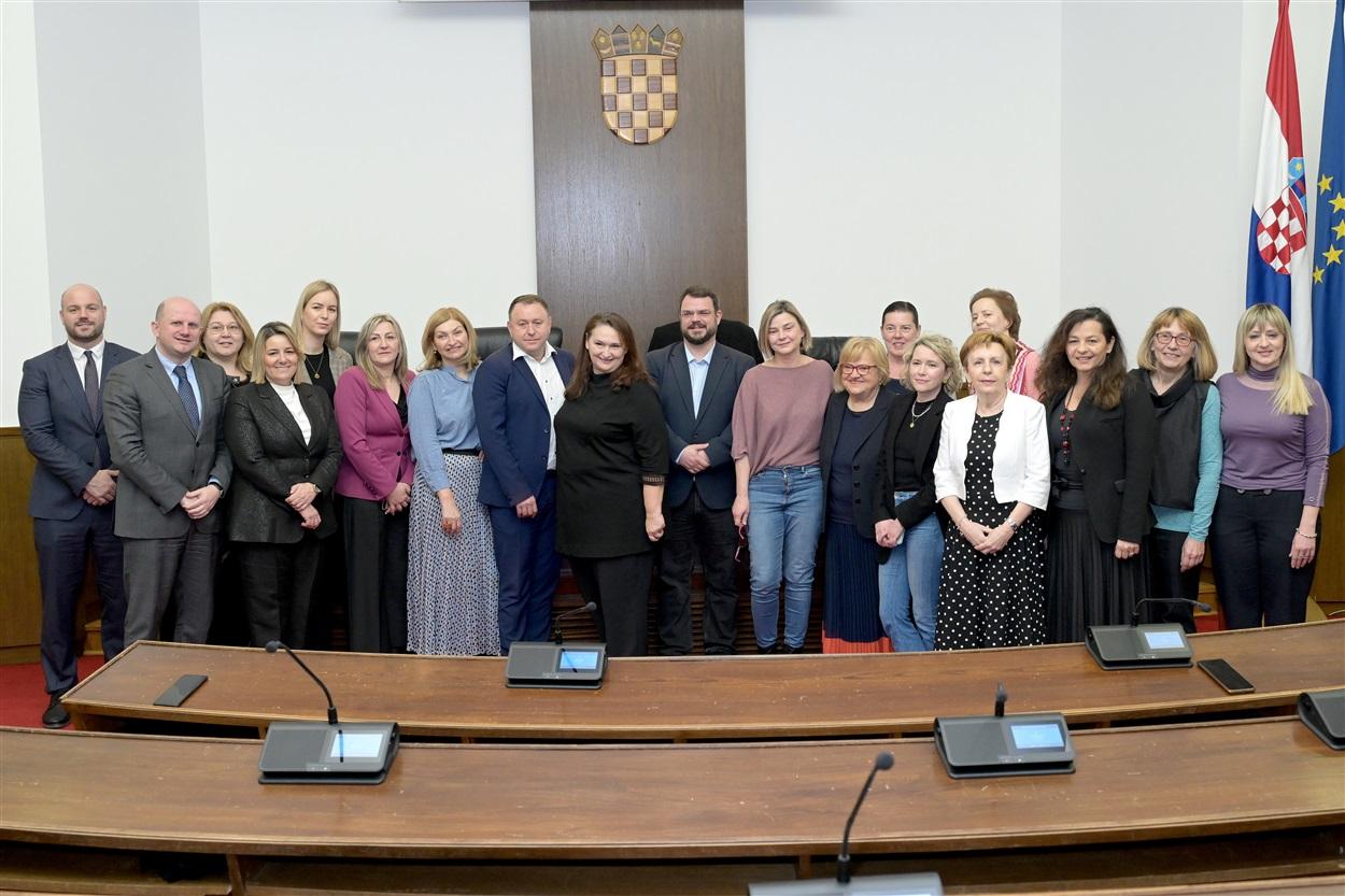 Na fotografiji je grupa ljudi koja stoji u prostoriji s državnim grbom i zastavama u pozadini. Ispred njih je dugi stol s mikrofonima. Ova postavka sugerira da se radi o službenom ili vladinom okruženju.