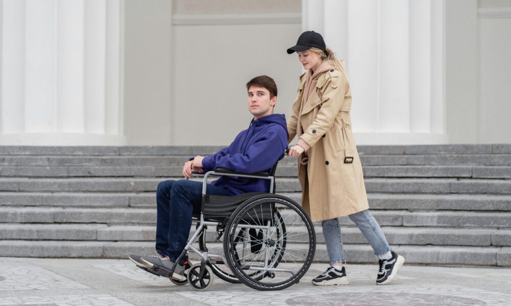 Žena u smeđoj jakni i crnoj kapi vozi čovjeka u kolicima. Čovjek je obučen u crne tenisice, crne hlače i plavu majicu. Prolaze pokraj zgrade s bijelim stupovima i stepenicama.