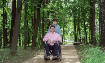 Na slici su dvije osobe na stazi u šumi. Jedna osoba je u invalidskim kolicima, a druga stoji iza nje. Osoba u invalidskim kolicima nosi ružičastu majicu, dok osoba koja gura kolica nosi plavu košulju. Okruženje je mirno i prirodno, s drvećem koje okružuje stazu.
