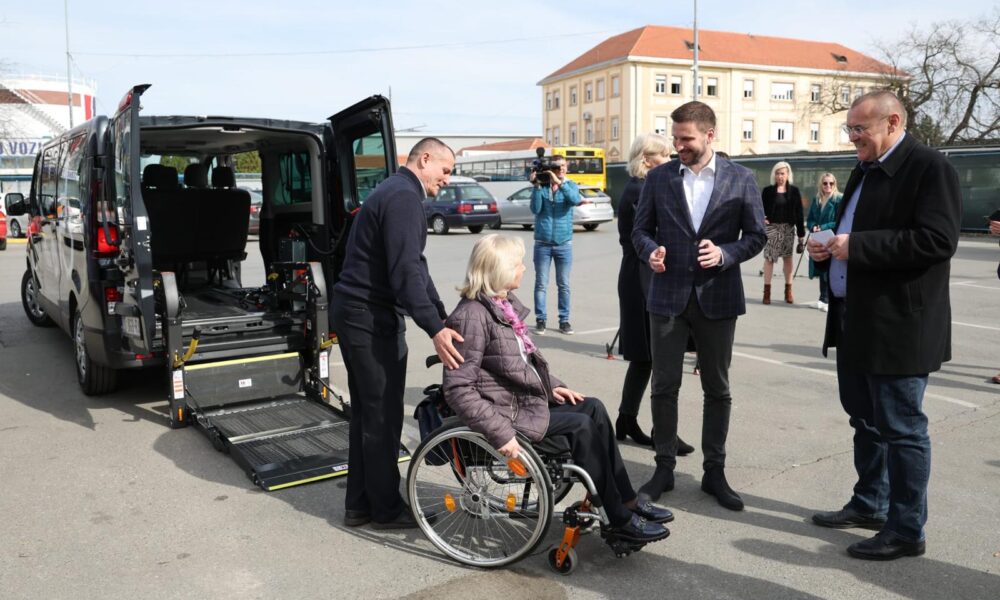 Na fotografiji je prikazana osoba u invalidskim kolicima koja komunicira s drugom osobom pored otvorenog kombija prilagođenog za prijevoz osoba s invaliditetom. Kombi je crne boje i ima otvorena stražnja vrata, otkrivajući rampu i specijaliziranu opremu unutar za osiguranje invalidskih kolica tijekom prijevoza. U pozadini su druge osobe i zgrada.