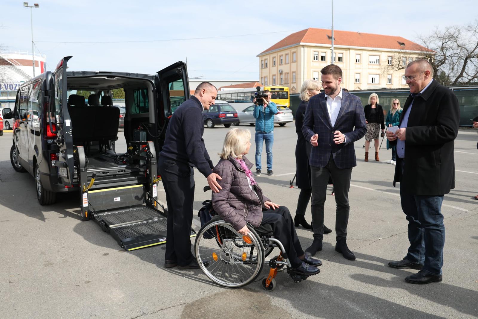 Na fotografiji je prikazana osoba u invalidskim kolicima koja komunicira s drugom osobom pored otvorenog kombija prilagođenog za prijevoz osoba s invaliditetom. Kombi je crne boje i ima otvorena stražnja vrata, otkrivajući rampu i specijaliziranu opremu unutar za osiguranje invalidskih kolica tijekom prijevoza. U pozadini su druge osobe i zgrada.