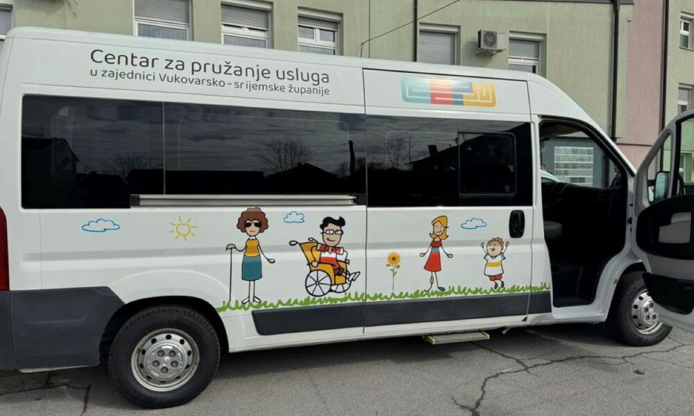 Na fotografiji je bijeli kombi parkiran na cesti. Na strani kombija je ilustracija koja prikazuje ljude različitih dobnih skupina, uključujući osobu u invalidskim kolicima. Iznad ilustracije piše “Centar za pružanje usluga u zajednici Vukovarsko-srijemske županije”. Kombijeva vrata su otvorena, a u pozadini se vidi zgrada. Ilustracija prikazuje sunčan dan s oblacima i travom.