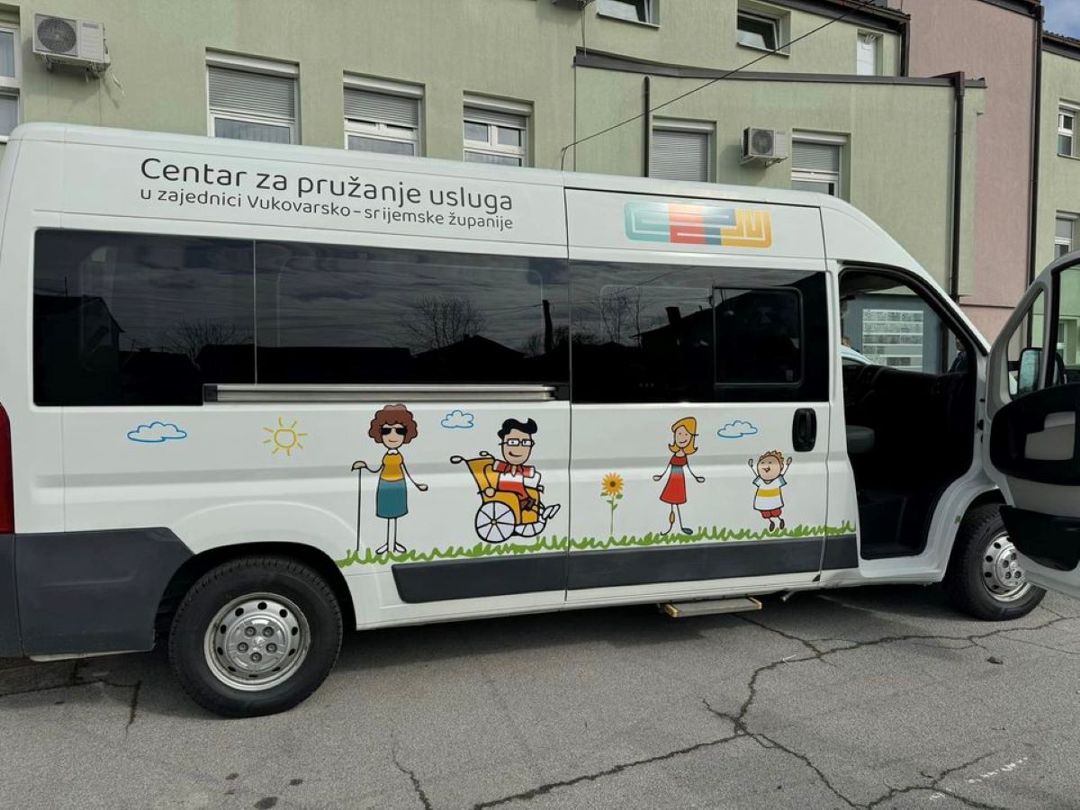 Na fotografiji je bijeli kombi parkiran na cesti. Na strani kombija je ilustracija koja prikazuje ljude različitih dobnih skupina, uključujući osobu u invalidskim kolicima. Iznad ilustracije piše “Centar za pružanje usluga u zajednici Vukovarsko-srijemske županije”. Kombijeva vrata su otvorena, a u pozadini se vidi zgrada. Ilustracija prikazuje sunčan dan s oblacima i travom.