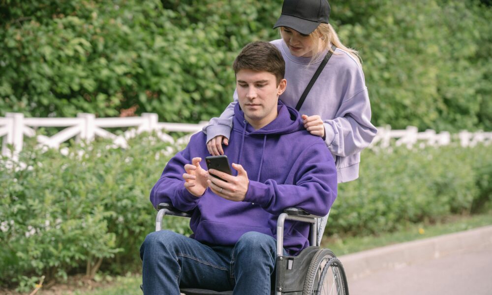 Na slici je osoba koja gura invalidska kolica u kojima sjedi druga osoba. Osoba u kolicima drži mobilni telefon u rukama. Oboje su odjeveni u pulovere i nalaze se na stazi pored zelene ograde i vegetacije.