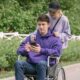 Na slici je osoba koja gura invalidska kolica u kojima sjedi druga osoba. Osoba u kolicima drži mobilni telefon u rukama. Oboje su odjeveni u pulovere i nalaze se na stazi pored zelene ograde i vegetacije.