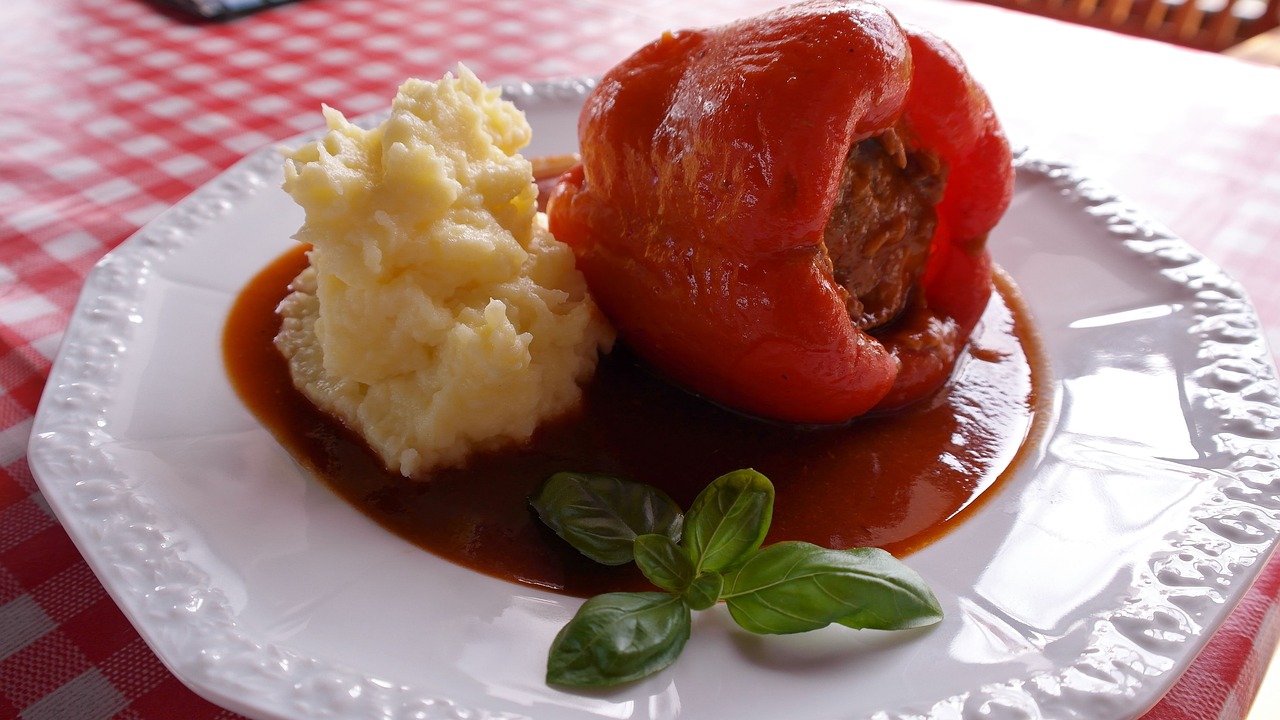 Na fotografiji je bijeli tanjur s ukusnim obrokom. Obrok se sastoji od punjene crvene paprike prelivene smeđim umakom i porcije pire krumpira. Paprika je ispunjena mesnom smjesom i prelivena umakom. Pored paprike i pire krumpira, na tanjuru je i nekoliko listova svježeg bosiljka koji dodaju estetski ugođaj jelu.