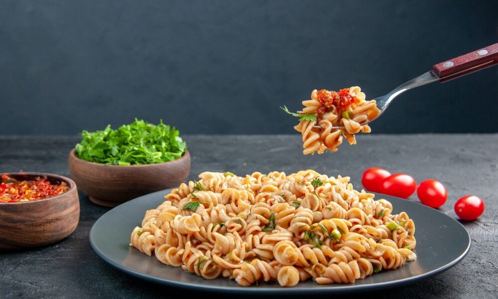 Na fotografiji je prikazana velika plava tanjura s tjesteninom posutom s nekim začinima i zelenilom, vjerojatno peršinom. Tjestenina izgleda ukusno i svježe pripremljena. Uz tanjur su smještene drvene zdjele, jedna s crvenim začinima i druga sa svježim zelenim salatama. Također, postoji vilica koja drži porciju tjestenine iznad tanjura, a u pozadini su crvene cherry rajčice.