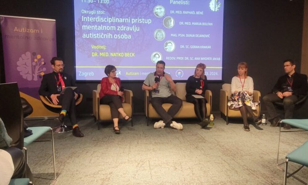 Fotografija prikazuje panel diskusiju u konferencijskoj dvorani. Na slici je pet osoba koje sjede na stolicama, a lica su im zamagljena. Iza njih je projekcijsko platno s informacijama o događaju napisanim na hrvatskom jeziku.