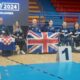 Na slici je skupina ljudi koji drže zastave Ujedinjenog Kraljevstva. i Republike Hrvatske Nalaze se u sportskoj dvorani, a iza njih je plavi zid s tekstom “ZAGREB/2024 BOCCIA CHALLENGER”. Osobe su obučene u tamnoplave trenirke i sjede u invalidskim kolicima, osim jedne osobe koja stoji. Ispred njih je tabla s brojem 1, ukazujući na to da su osvajači medalja.