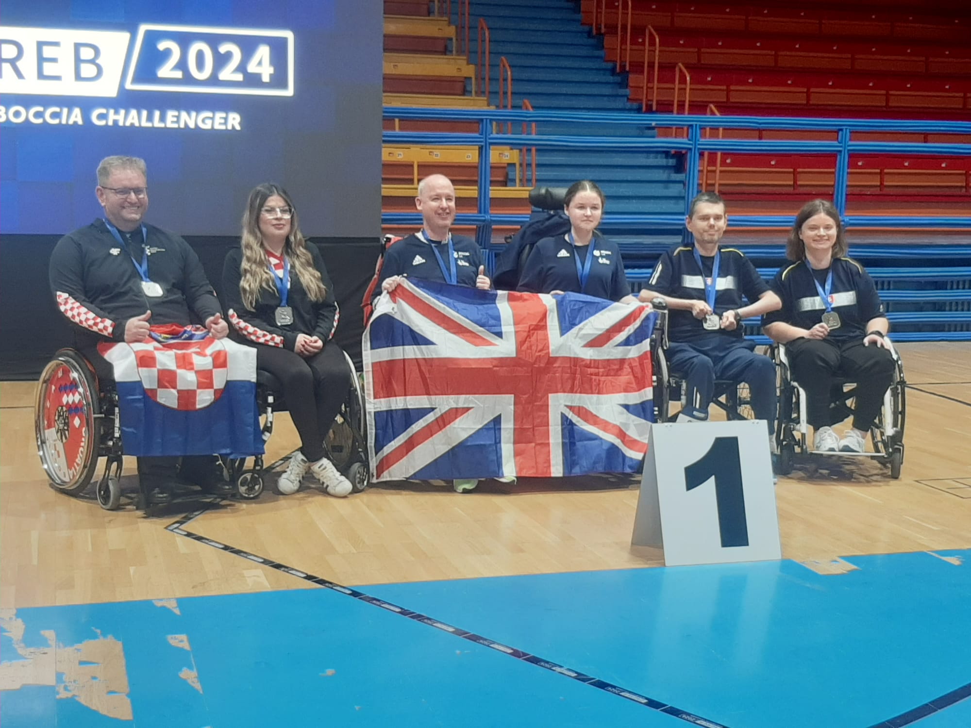 Na slici je skupina ljudi koji drže zastave Ujedinjenog Kraljevstva. i Republike Hrvatske Nalaze se u sportskoj dvorani, a iza njih je plavi zid s tekstom “ZAGREB/2024 BOCCIA CHALLENGER”. Osobe su obučene u tamnoplave trenirke i sjede u invalidskim kolicima, osim jedne osobe koja stoji. Ispred njih je tabla s brojem 1, ukazujući na to da su osvajači medalja.