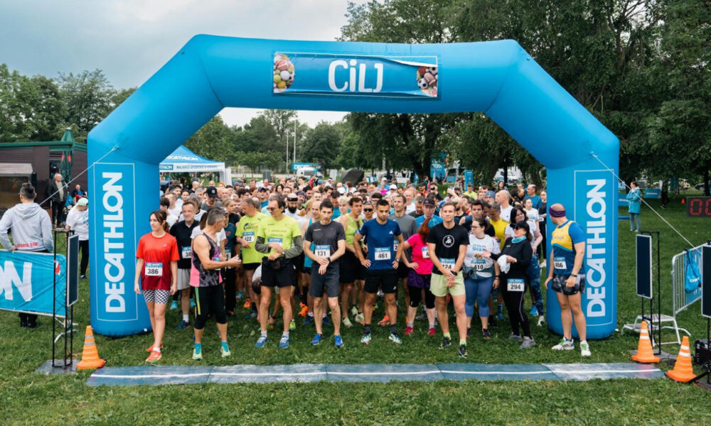 Na slici je skupina ljudi spremnih za početak utrke. Stoje ispod velikog plavog napuhanog luka s natpisima “DECATHLON” i “CIlJ”. Sudionici su odjeveni u različitu sportsku odjeću, uključujući kratke hlače, majice i potkošulje. Na mnogima su vidljivi brojevi za utrku, što ukazuje na to da su dio organiziranog događaja. Atmosfera izgleda živahno i energično s zelenim drvećem u pozadini, što sugerira da se radi o vanjskom prostoru, vjerojatno u parku. Na lijevoj strani je osoba koja čini se upravlja ili promatra događaj, stoji pored transparenta također označenog s “DECATHLON”.