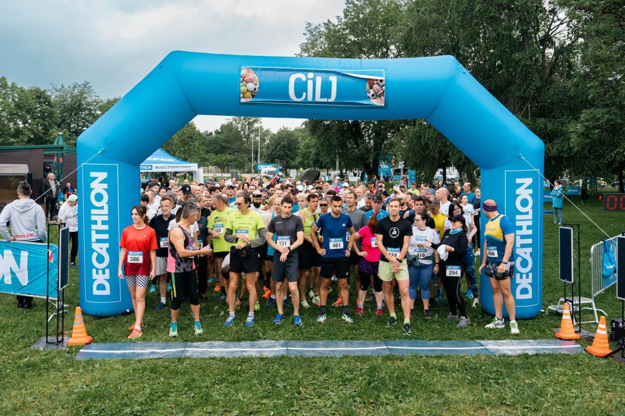 Na slici je skupina ljudi spremnih za početak utrke. Stoje ispod velikog plavog napuhanog luka s natpisima “DECATHLON” i “CIlJ”. Sudionici su odjeveni u različitu sportsku odjeću, uključujući kratke hlače, majice i potkošulje. Na mnogima su vidljivi brojevi za utrku, što ukazuje na to da su dio organiziranog događaja. Atmosfera izgleda živahno i energično s zelenim drvećem u pozadini, što sugerira da se radi o vanjskom prostoru, vjerojatno u parku. Na lijevoj strani je osoba koja čini se upravlja ili promatra događaj, stoji pored transparenta također označenog s “DECATHLON”.