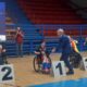 Na slici je ceremonija dodjele medalja u sportskoj dvorani. Tri osobe u invalidskim kolicima su na podiju označenom brojevima 1, 2 i 3. Osoba koja je osvojila prvo mjesto nosi crvenu majicu i drži zastavu na koljenima, a osoba koja dodjeljuje nagrade nosi plavo odijelo. Na ekranu u pozadini prikazane su informacije o natjecatelju.