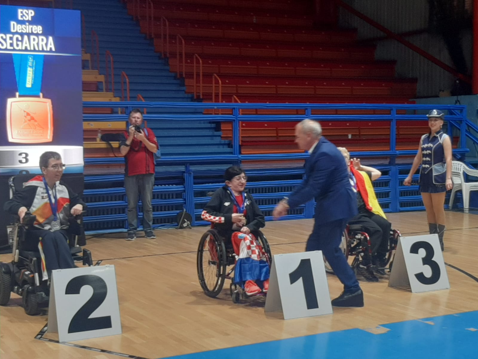 Na slici je ceremonija dodjele medalja u sportskoj dvorani. Tri osobe u invalidskim kolicima su na podiju označenom brojevima 1, 2 i 3. Osoba koja je osvojila prvo mjesto nosi crvenu majicu i drži zastavu na koljenima, a osoba koja dodjeljuje nagrade nosi plavo odijelo. Na ekranu u pozadini prikazane su informacije o natjecatelju.