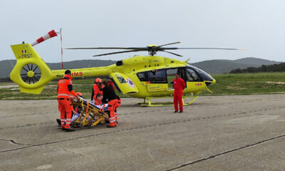 Ova slika prikazuje trenutak kada hitni medicinski tim u narančastim odijelima pruža pomoć pacijentu na nosilima, dok je žuti medicinski helikopter spreman za prijevoz.