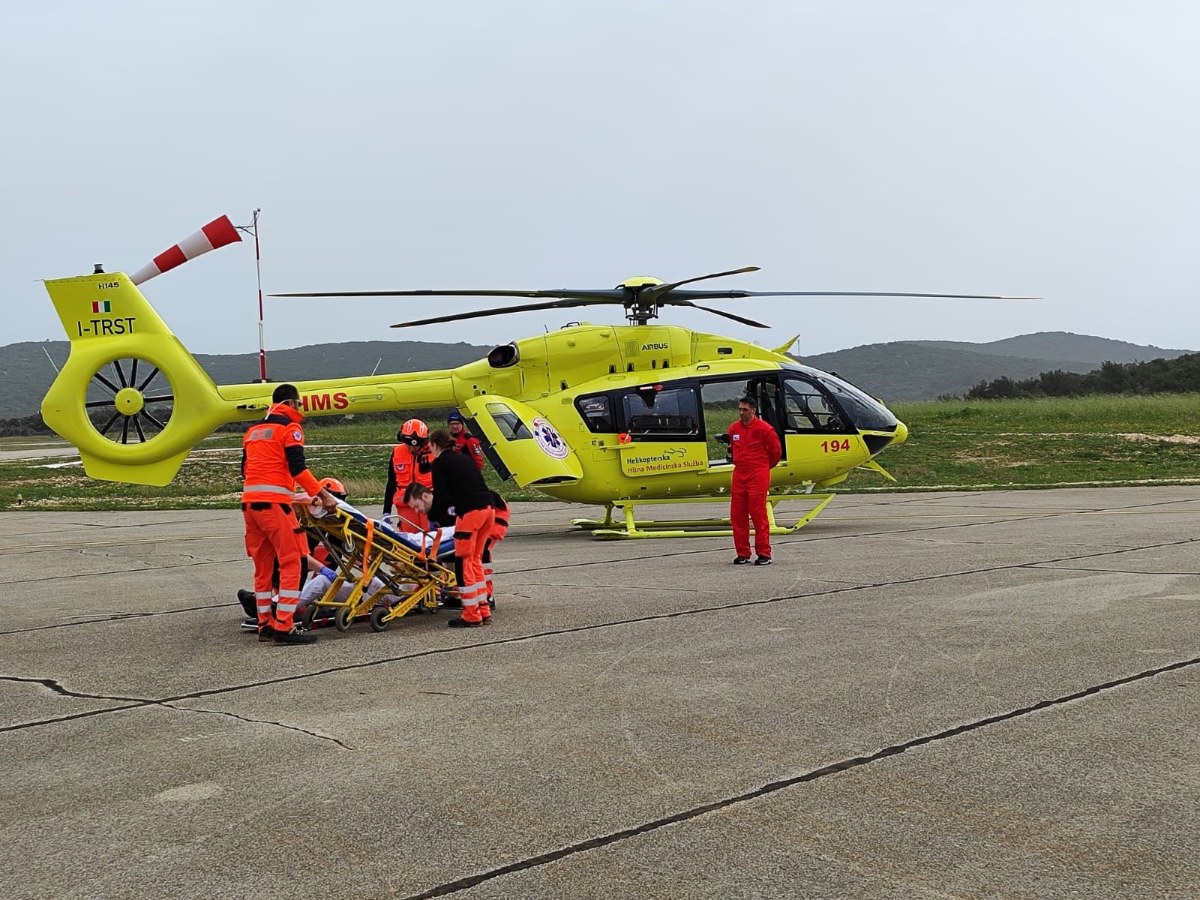 Ova slika prikazuje trenutak kada hitni medicinski tim u narančastim odijelima pruža pomoć pacijentu na nosilima, dok je žuti medicinski helikopter spreman za prijevoz.