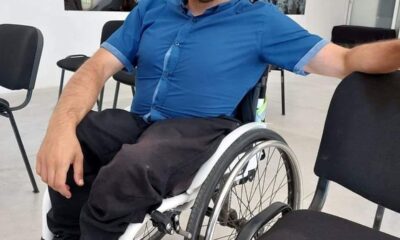 Na fotografiji je osoba u invalidskim kolicima koja nosi plavu košulju i crne hlače. Lice osobe nije vidljivo. Osoba se nalazi u unutarnjem prostoru s bijelim zidovima na kojima su obješene crno-bijele slike. U pozadini se mogu primijetiti crne stolice. Pod je svijetle boje, ali detalji nisu jasno vidljivi.