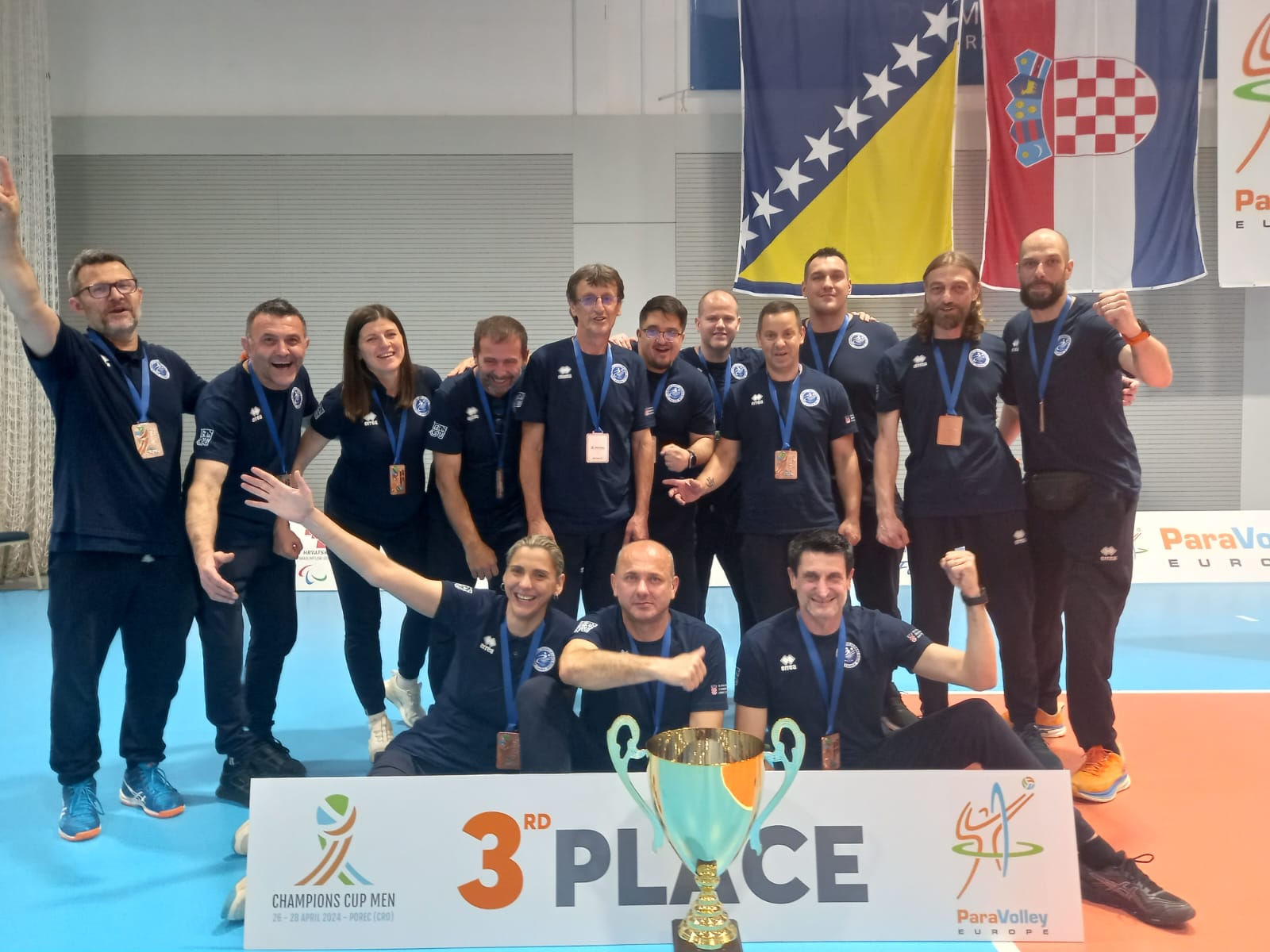 Na slici je grupa ljudi u sportskim uniformama koji poziraju s velikim peharom. Iza njih su zastave, a ispred njih je natpis “3RD PLACE”. Slika je očito sa nekog sportskog događanja gdje su osvojili treće mjesto. Pozadina sugerira da se radi o natjecanju u odbojci za osobe s invaliditetom, s obzirom na tekst “ParaVolley” koji se može vidjeti na slici.