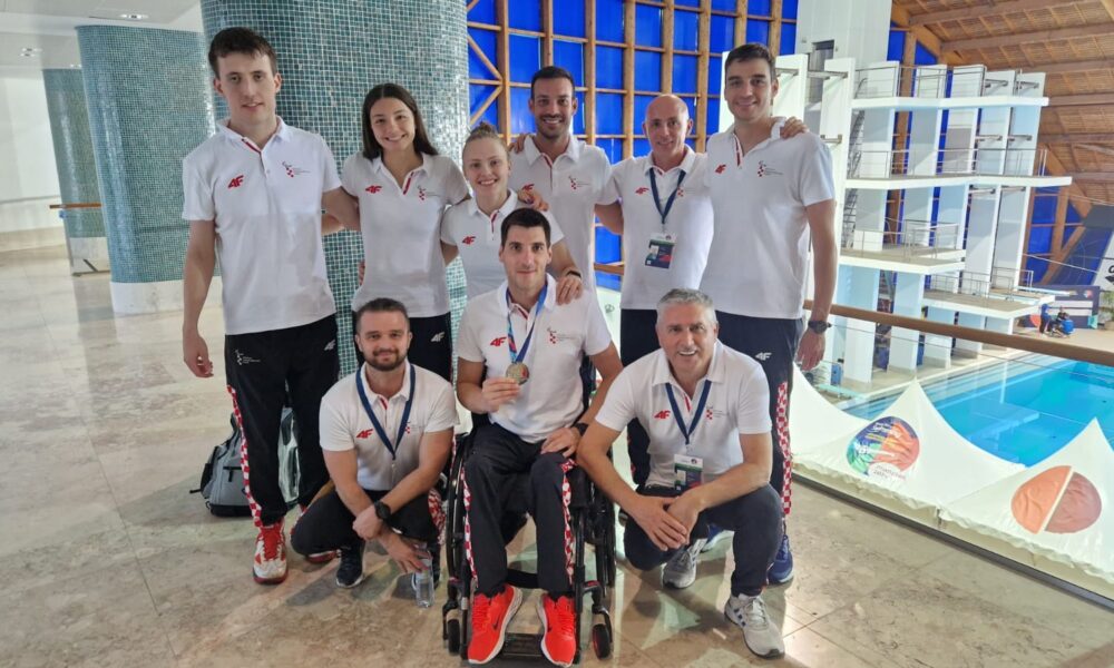 Na slici su osobe u bijelim majicama, jedna osoba sjedi u invalidskim kolicima i drži medalju u ruci. Ljudi stoje pokraj bazena što upućuje da se radi o plivačkom natjecanju.